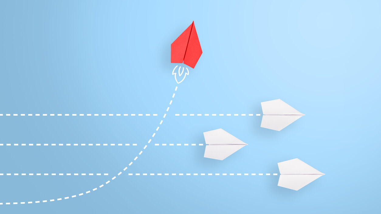 Weiße Papierflieger fliegen in eine Richtung, roter Papierflieger fliegt nach oben. Bild symbolisiert Brand Driven Transformation
